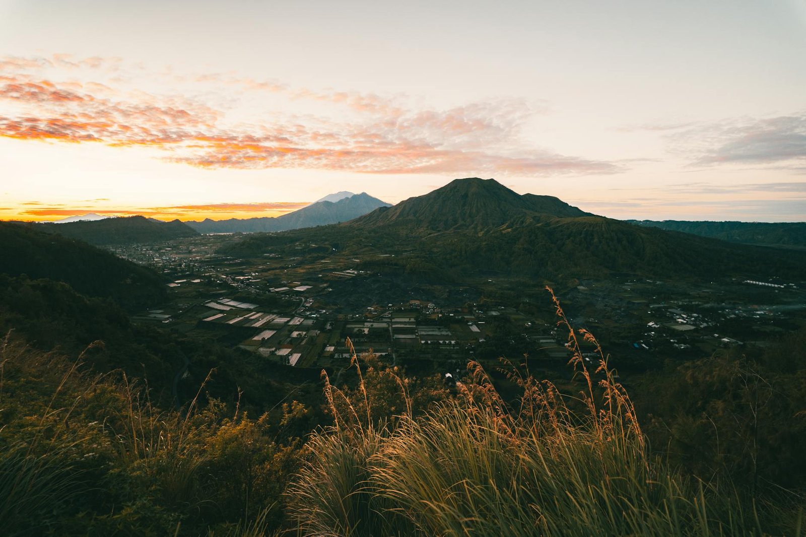 Sunrise view of Mount Batur and surrounding landscape in Bali, Indonesia, featuring lush vegetation and distant mountains.